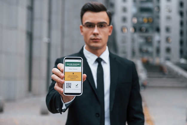 Holding phone with vaccination certificate Businessman in black suit and tie is outdoors in the city