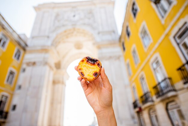 Tenendo la pasticceria portoghese della crostata dell'uovo chiamata pastel de nata all'aperto sullo sfondo dell'arco trionfale a lisbona