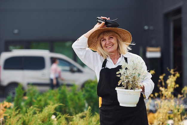 Holding pot in hands Senior woman is in the mini garden at daytime Building exterior behind