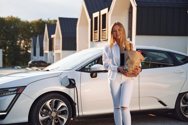 買い物袋を持っている白い服を着た若い女性が昼間に電気自動車に乗っている