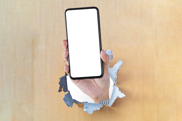 Holding smartphone with blank screen in hand through hole on brown cardboard background