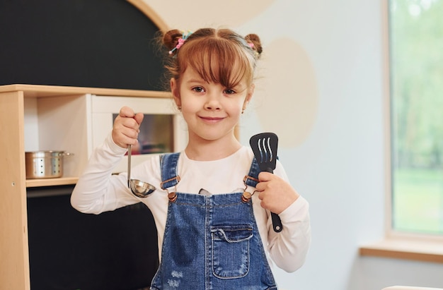 道具を手に持って見せてくれるカジュアルな服装の女の子がキッチンでおもちゃで遊んで楽しんでいます