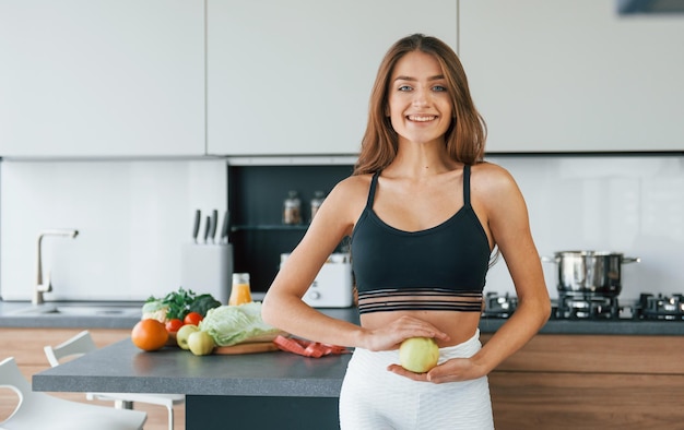 Holds apple against belly Young european woman is indoors at kitchen indoors with healthy food