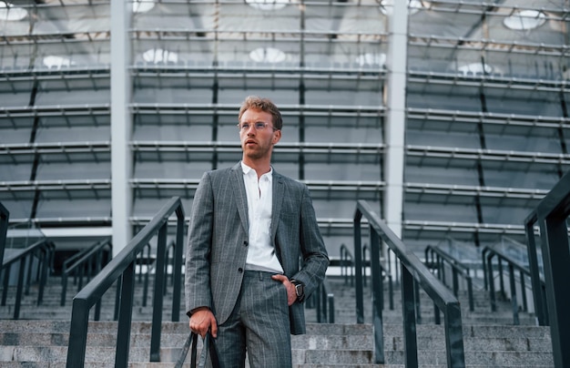 Holds bag in hands Young businessman in grey formal wear is outdoors in the city