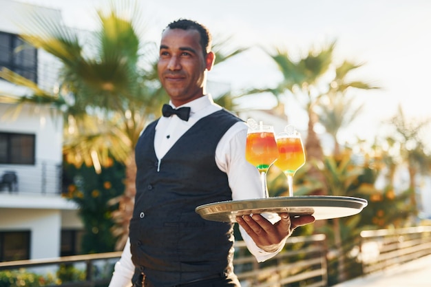 Holds cocktails Black waiter in formal clothes is at his work outdoors at sunny daytime