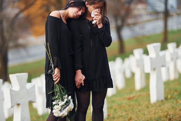꽃을 들고 검은 옷을 입은 두 젊은 여성이 많은 흰색 십자가가 있는 묘지를 방문합니다. 장례식과 죽음의 개념