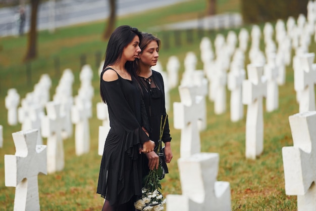 Holds flowers two young women in black clothes visiting\
cemetery with many white crosses conception of funeral and\
death