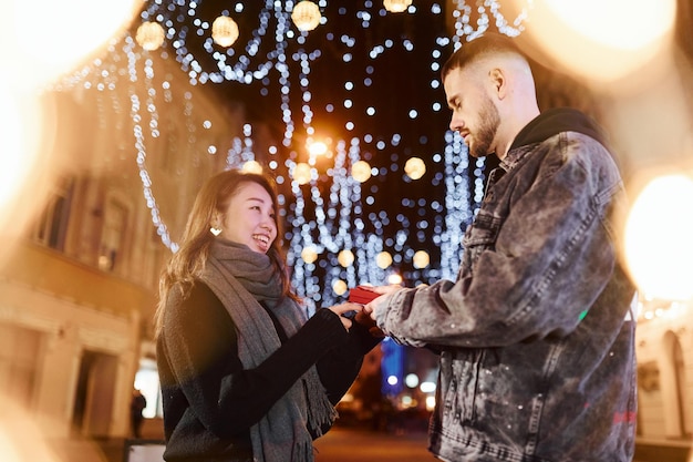 Holds gift box Happy multiracial couple together outdoors in the city celebrating New year