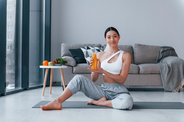 Holds glass with orange juice in hands Young woman with slim body shape in sportswear have fitness day indoors at home