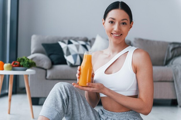 Holds glass with orange juice in hands Young woman with slim body shape in sportswear have fitness day indoors at home