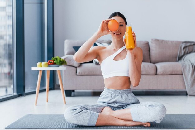 Holds glass with orange juice in hands Young woman with slim body shape in sportswear have fitness day indoors at home