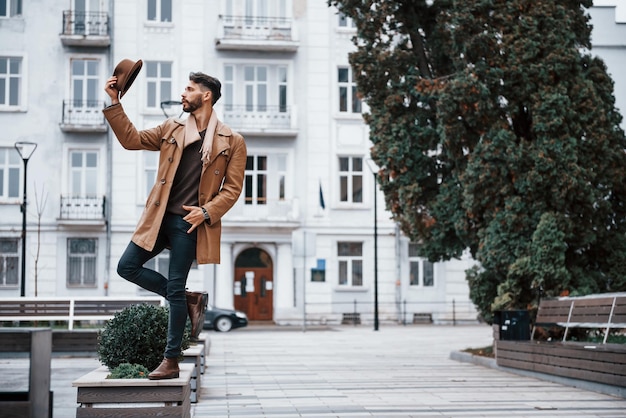 Holds hat and posing Young male model in fashionable clothes is outdoors in the city at daytime