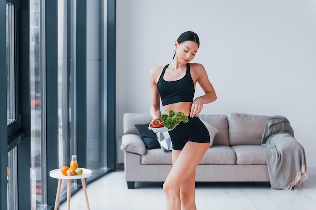 Holds healthy food in hands Young woman with slim body shape in black sportswear have fitness day indoors at home