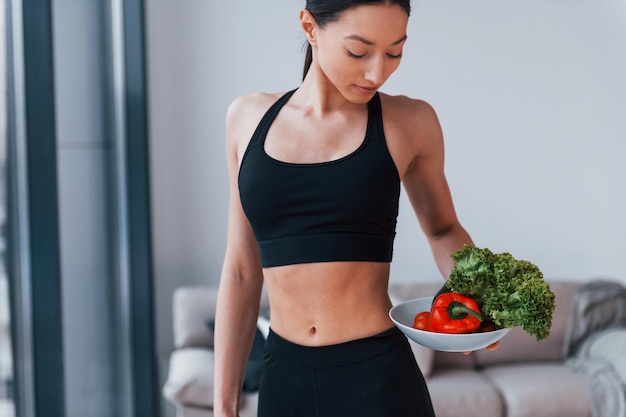 Holds healthy food in hands Young woman with slim body shape in black sportswear have fitness day indoors at home