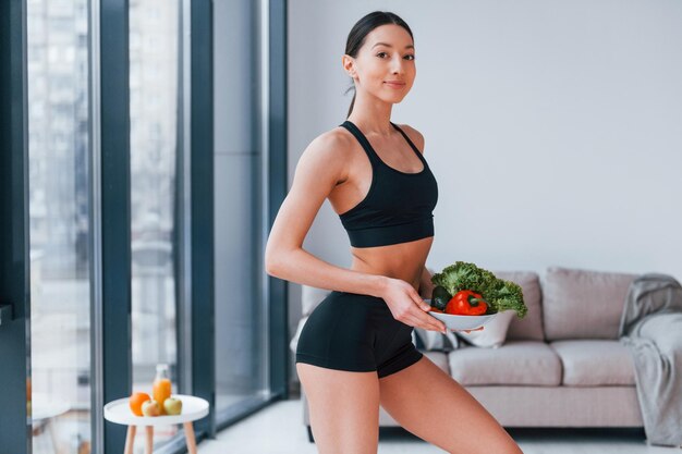 Holds healthy food in hands Young woman with slim body shape in black sportswear have fitness day indoors at home