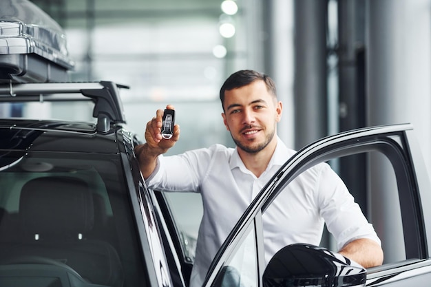 Holds keys Young man in white shirt is indoors with modern new automobile