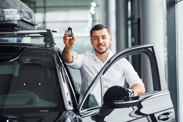 Holds keys Young man in white shirt is indoors with modern new automobile