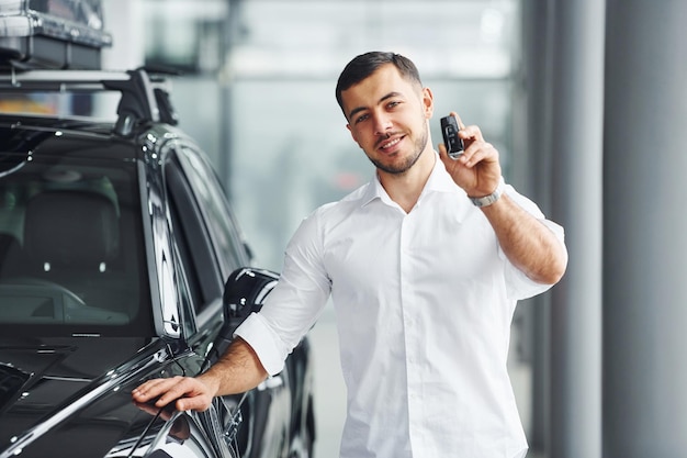 Holds keys Young man in white shirt is indoors with modern new automobile