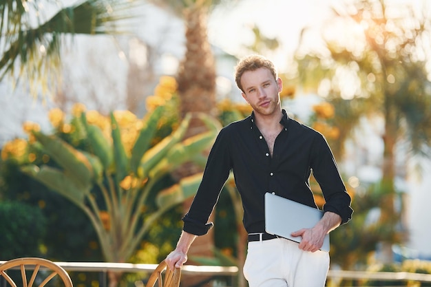 Holds laptop Young man is outdoors at sunny daytime Concept of vacation