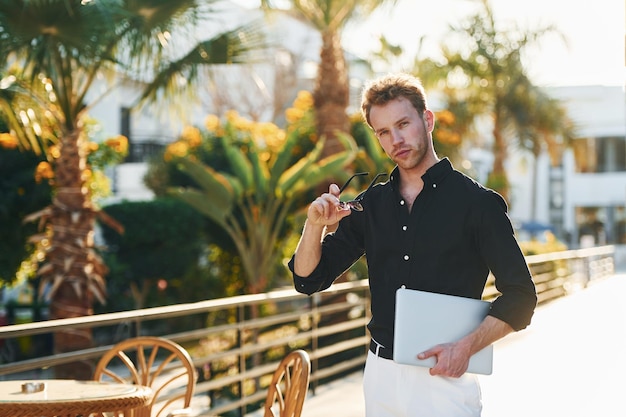 Holds laptop Young man is outdoors at sunny daytime Concept of vacation