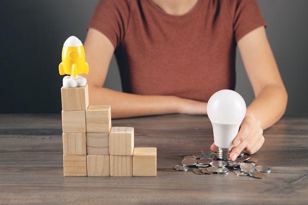 Photo holds a light bulb near a coin and a rocket stands on a ladder of cubes