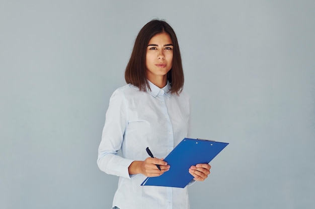 Holds notepad and pen Young modern woman in white shirt standing inside of the studio