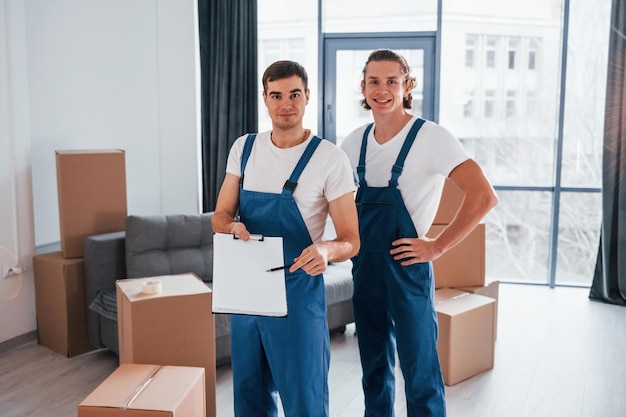 Holds notepad with document Two young movers in blue uniform working indoors in the room