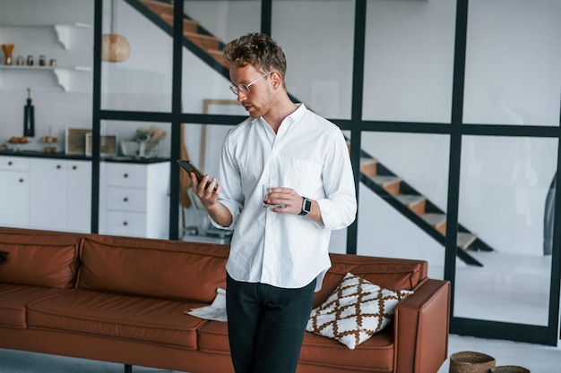 Holds phone and glass Caucasian young guy in elegant white shirt indoors at home