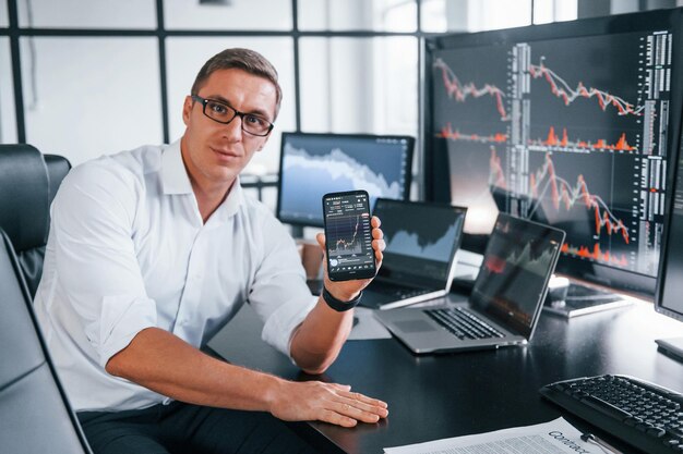 holds phone young male stock brocker works indoors in the office many of displays
