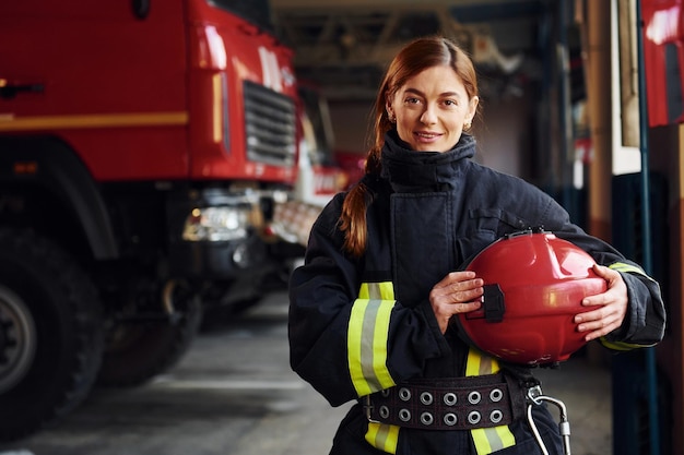 Holds red hat in hands Female firefighter in protective uniform standing near truck