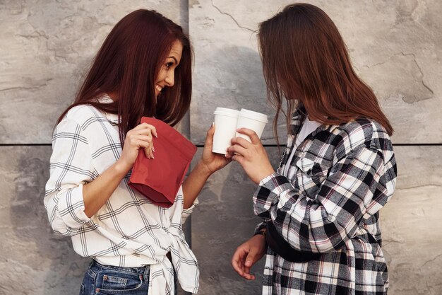 Holds red package of coffee beautiful cheerful friends or\
lesbian couple together near wall outdoors at daytime