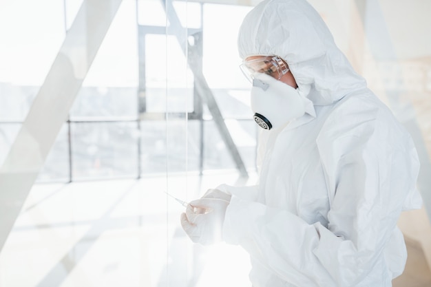 Holds syringe with medicine. Female doctor scientist in lab coat, defensive eyewear and mask standing indoors