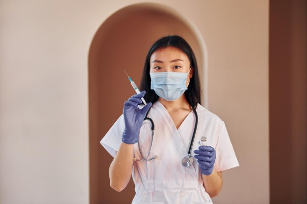 Holds vaccine and syringe Young serious asian woman standing indoors