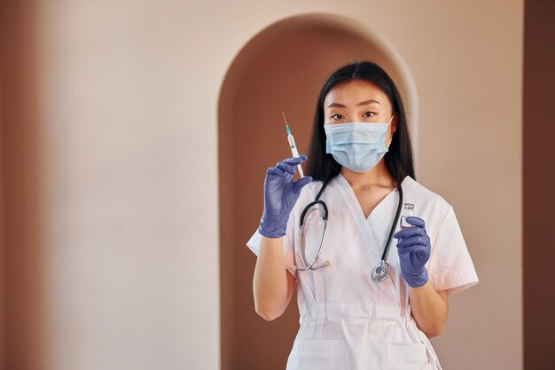 Holds vaccine and syringe Young serious asian woman standing indoors