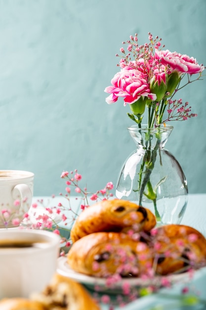 ターコイズブルーの表面にコーヒー、ミニ焼きたてのクロワッサンチョコレートパン、カーネーションの花を添えたホリデーブレイク。コピースペース