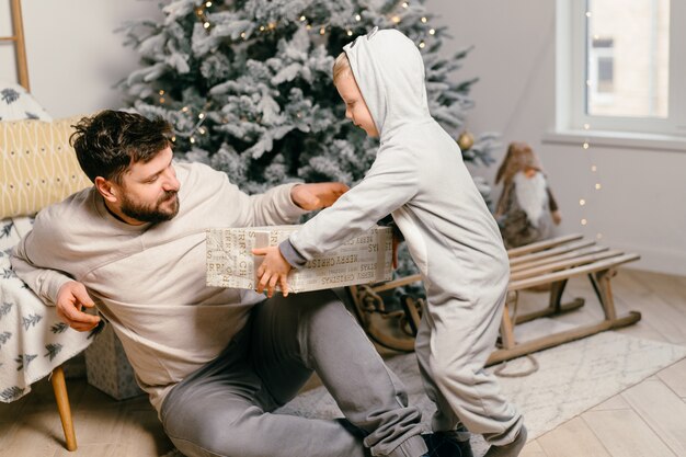 Holiday Christmas Handsome father playing with small cute son near decorated New year tree at home Family tradition boy gives a gift to his father