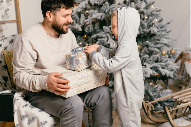 休日のクリスマス家で飾られた新年の木の近くで小さなかわいい息子と遊ぶハンサムな父家族の伝統的な男の子は彼の父に贈り物をします