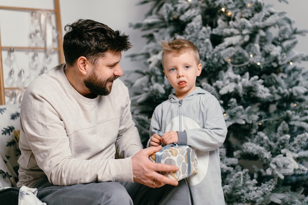 休日のクリスマス家で飾られた新年の木の近くで小さなかわいい息子と遊ぶハンサムな父家族の伝統の父は彼の小さな男の子に贈り物をします