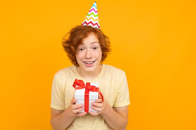 Holiday concept. happy surprised red-haired boy with a holiday cap on his head with a gift in his hands on an orange background 