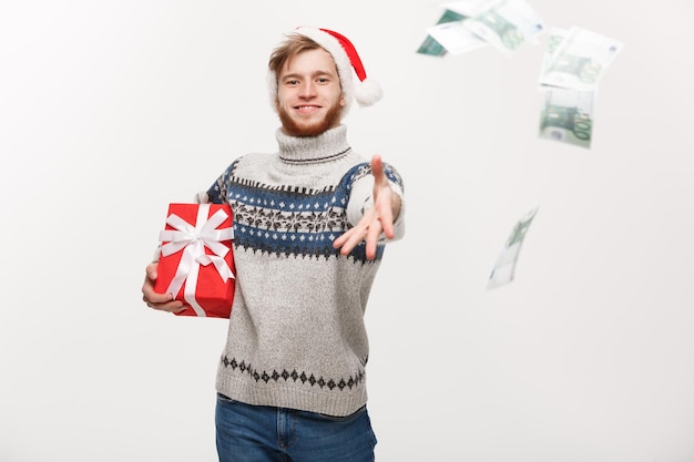Концепция праздника Молодой бородатый мужчина держит рождественский подарок и бросает деньги в камеру