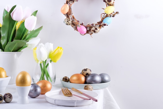 Holiday congratulation composition of served table with handmade painted eggs, baked cookies, fresh spring tulips flowers and festive wreath on a light grey wall. Happy Easter concept.