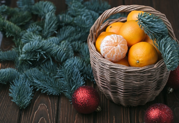 Foto contenuto festivo, mandarini, mandarino sbucciato in un cesto di vimini, vintage, ramo di abete, palle rosse dell'albero di natale, sfondo marrone scuro, vista dall'alto