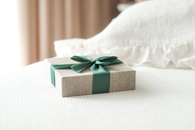 휴일 선물 및 고급 온라인 쇼핑 배달은 침실의 세련된 시골 스타일의 침대에 녹색 리본이 달린 린넨 선물 상자를 포장합니다.