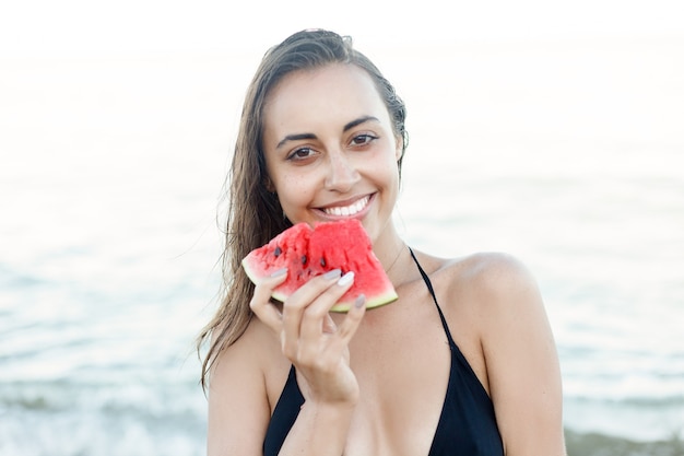 Holiday, resort, tourism concept - Summer vacation - young girl eating fresh watermelon on sandy beach. young beautiful woman eats watermelon on the beach at hot summer day.