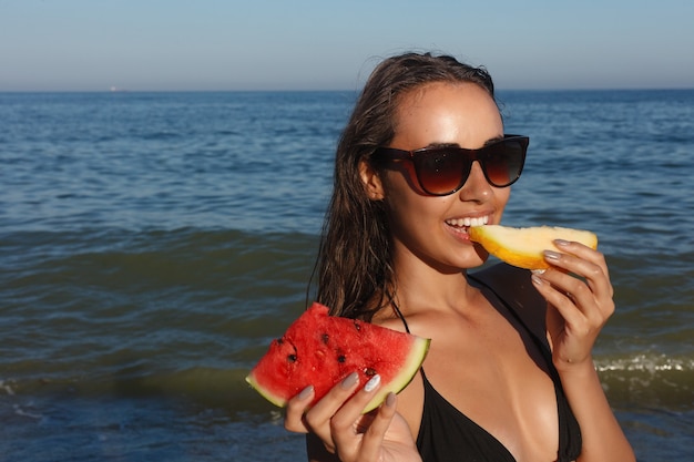 休日、リゾート、観光のコンセプト-夏休み-砂浜で新鮮なスイカを食べる少女。若い美しい女性は暑い夏の日にビーチでスイカを食べます。