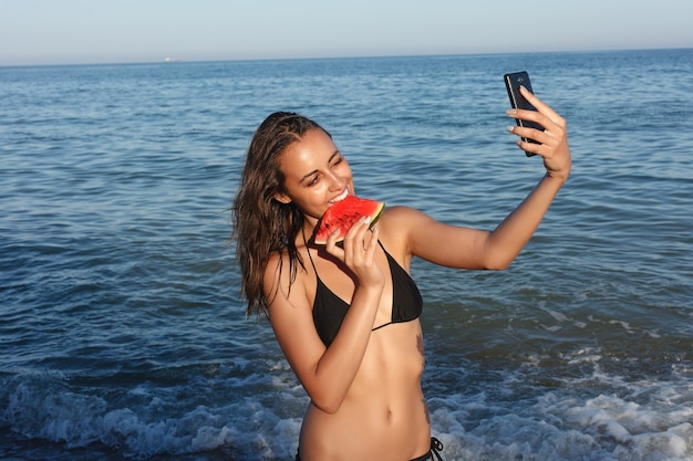 Foto vacanze, resort, concetto di turismo - vacanze estive - ragazza che mangia anguria fresca sulla spiaggia sabbiosa. giovane bella donna mangia anguria sulla spiaggia al caldo giorno d'estate.