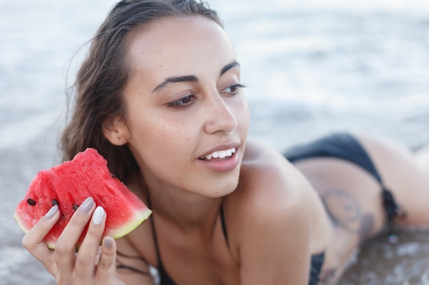 Vacanze, resort, concetto di turismo - vacanze estive - ragazza che mangia anguria fresca sulla spiaggia sabbiosa. giovane bella donna mangia anguria sulla spiaggia al caldo giorno d'estate.