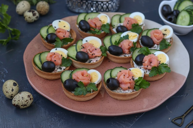 Holiday tartlets with cheese, salmon, black olives, quail eggs and cucumbers on dark surface, horizontal orientation, closeup