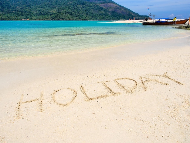 Holiday text on white sand beach with blue sea and mountain background