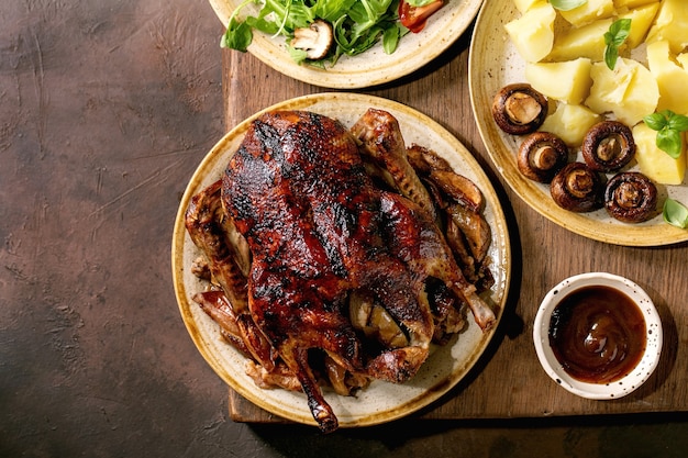 Holiday Thanksgiving table with classic dishes roasted glazed duck with apples, boiled potatoes, green salad and sauce on dark wooden table with autumn decor. Flat lay, copy space